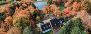 Aerial view of a large manor house at Edson Hill - Stowe Vermont Inn, surrounded by trees with vibrant autumn foliage in shades of orange, red, and green, with a small pond visible in the background.