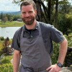 Executive Chef Matt Hiebsch stands outdoors in a gray apron and short-sleeve shirt, smiling amid the lush greenery, pond, and hills of Edson Hill.