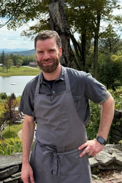 Executive Chef Matt Hiebsch stands outdoors in a gray apron and short-sleeve shirt, smiling amid the lush greenery, pond, and hills of Edson Hill.