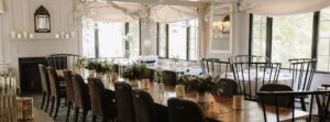 Elegant dining room at Edson Hill - Stowe Vermont Inn with a long wooden table, black high-back chairs, floral centerpieces, and candles. Large windows let in natural light while glass orbs hang from white decorative branches above.