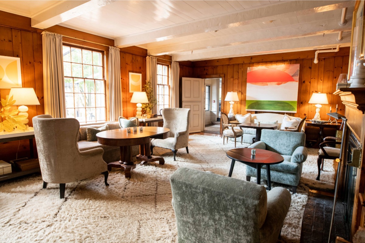 Cozy, sunlit living room with wood-paneled walls,  white cieling with hand-hewn beams from Ethan Allen's homestead, plush armchairs, round tables, lamps, abstract art, and large windows—perfect for intimate gaterings—features a soft, cream-colored rug covering the floor.