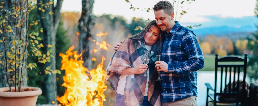 A couple stands outdoors by a fire pit at Edson Hill - Stowe Vermont Inn, embracing and smiling with glasses of wine. Autumn trees and a tranquil pond provide a stunning backdrop.
