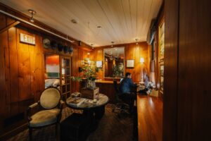 A cozy wooden front office for guest reception with warm lighting, featuring a round table with plants, vintage chairs, and wall art. A person in a blue checked shirt works by the window, while an illuminated exit sign hangs above the door.