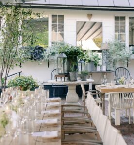 Outdoor dining area with teak tables, white chairs, and elegant table settings. Lush green plants and white flowers decorate the space, creating a cozy, garden-like Event Gallery perfect for gatherings.