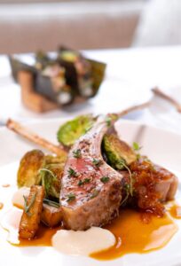 A plated gourmet dish at Edson Hill - Stowe Vermont Inn features rack of lamb, garnished with herbs, served with roasted vegetables, creamy white sauce, and a rich brown sauce, with another enticing dish blurred in the background.