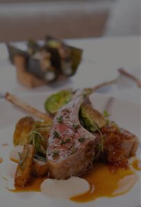 A plated gourmet dish at Edson Hill - Stowe Vermont Inn features lamb chops garnished with herbs, roasted vegetables, and a rich brown sauce, artfully arranged on a white plate. Another elegant dish is softly blurred in the background.