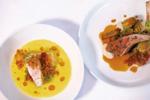 Two white plates with gourmet dishes by Edson Hill's new executive chef in Stowe, Vermont: one features fish in a yellow sauce with red oil and crumbs, the other offers sliced lamb with creamy sauce, fried Brussels sprouts, and brown gravy. Both are artfully arranged.