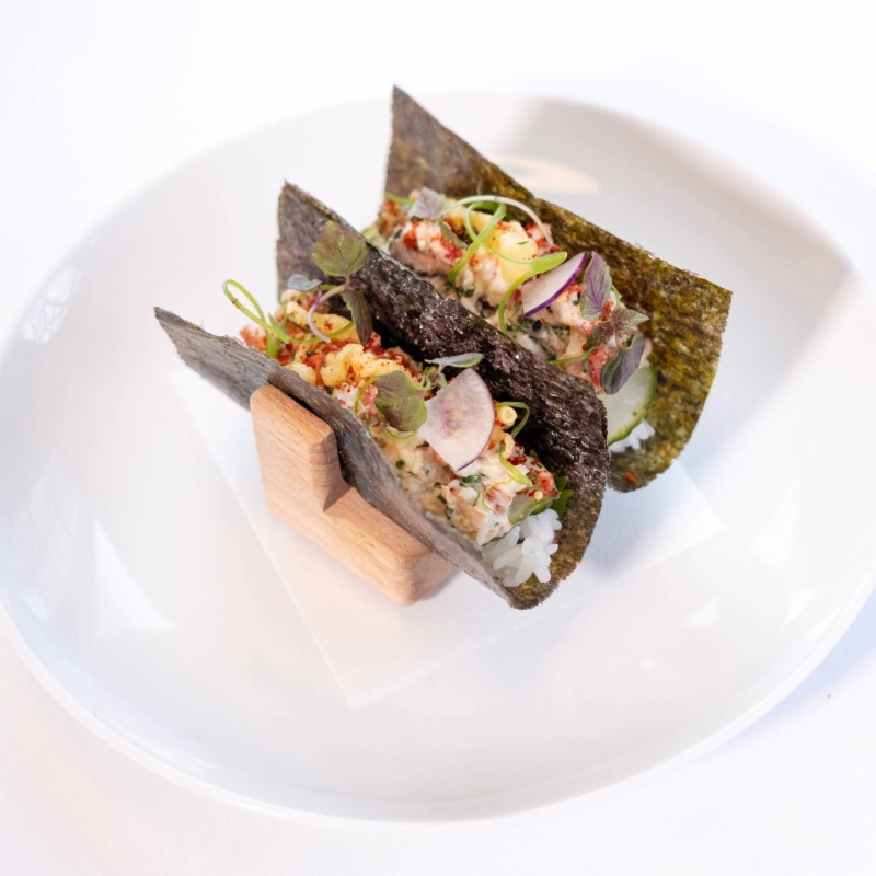 Two hand rolls with seaweed wrappers, filled with rice, vegetables, microgreens, and thinly sliced radish, are displayed upright in a wooden holder on a white plate—a perfect bite for Fine Dining in Stowe VT.
