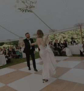 A bride and groom dance together on a checkered floor under a white tent, surrounded by smiling guests. This heartwarming moment is a highlight in our Event Gallery.