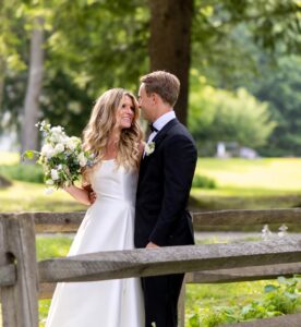 A bride in a white dress holding a bouquet and a groom in a black suit stand smiling at each other outdoors by a wooden fence, surrounded by greenery and trees on a sunny day—perfect for an Event Gallery highlight.