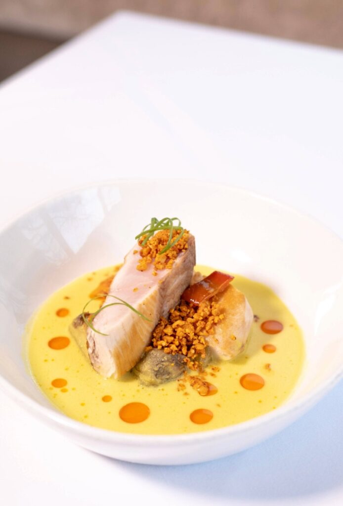 A white bowl with pieces of roasted pork belly on yellow creamy sauce, garnished with crispy bits, green herbs, and drops of orange oil—an artful dish that reflects the essence of casual fine dining on a white tablecloth.