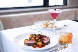 A white table set for casual fine dining features a plate of sliced beef, roasted vegetables, and sauce, paired with an old fashioned cocktail. In the background, another dish and a pink cocktail add to the inviting atmosphere.