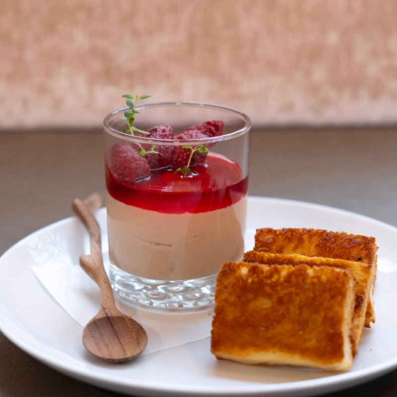 A glass cup with creamy dessert topped with red berry sauce and strawberries, garnished with herbs, sits on a white plate with three golden toast slices and a small wooden spoon—a sweet highlight of our Farm to Table Menu Vermont.