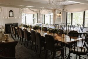 A long wooden dining table with dark chairs is set in an elegant, sunlit room decorated with candles, flowers, and hanging branches—perfect for a casual fine dining experience. Large windows let in natural light, with a white fireplace in the background.