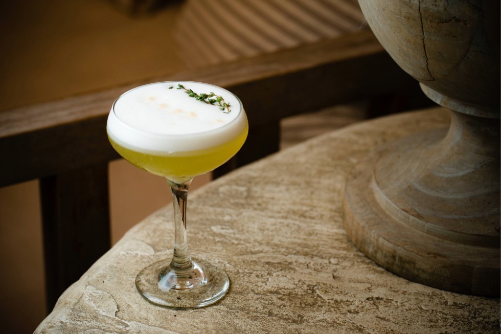 A cocktail in a coupe glass with a frothy white top, garnished with a small sprig of green herbs, sits on a rustic stone table near a decorative column—a perfect touch for any casual fine dining experience.