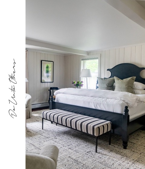 A bright, cozy bedroom with a dark wood bed, white bedding, gray pillows, a striped bench at the foot, a textured rug, and light walls evokes the inviting charm of Vermont luxury hotel accommodations. "Do Unto Others" appears on the left.