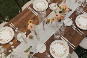 A wooden table set for a wedding reception at a Stowe wedding venue features floral-patterned plates, menus, clear glasses, silver cutlery, and small vases of pastel flowers on a white runner, with fruit scattered among the arrangements.