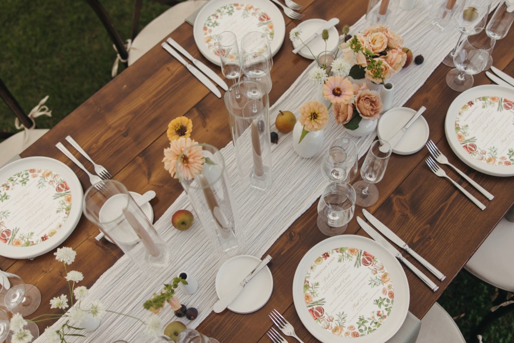 A wooden table set for a wedding reception at a Stowe wedding venue features floral-patterned plates, menus, clear glasses, silver cutlery, and small vases of pastel flowers on a white runner, with fruit scattered among the arrangements.