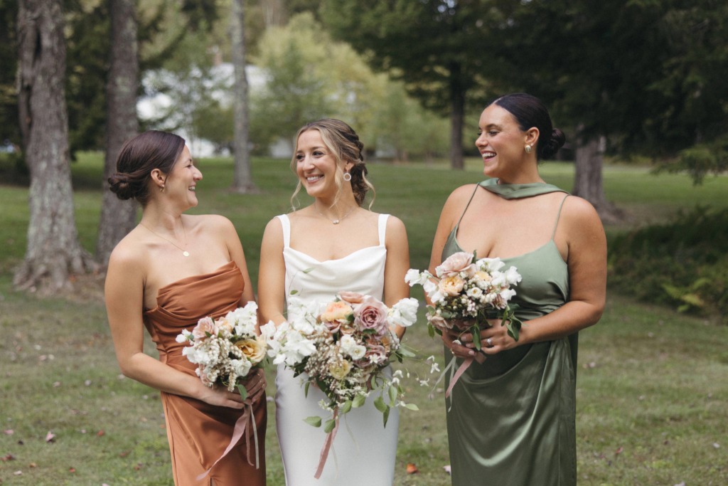 Three women in satin dresses stand outdoors at a Stowe wedding venue, smiling and holding flower bouquets. The woman in the center wears white, while the others wear rust and green. Trees and grass fill the background on a bright day.