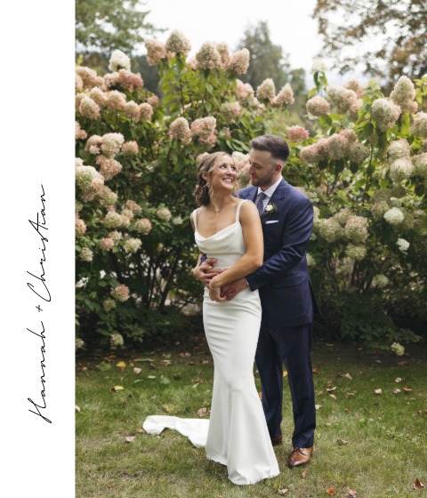 A bride in a white gown and groom in a navy suit stand closely together, smiling lovingly, surrounded by blooming hydrangea bushes at a picturesque Stowe wedding venue. Handwritten text on the side reads, Hannah + Christian.