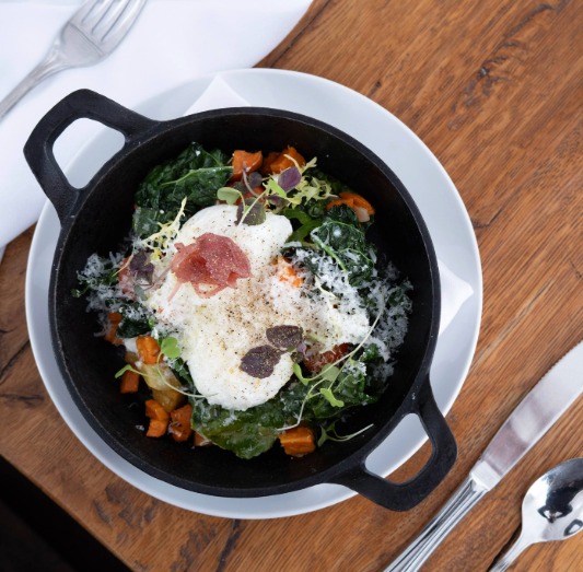 A cast iron skillet filled with cooked greens, diced vegetables, a poached egg topped with grated cheese and a small slice of cured meat—perfect for a Mother's Day Brunch in Stowe—served on a white plate with cutlery on a wooden table.