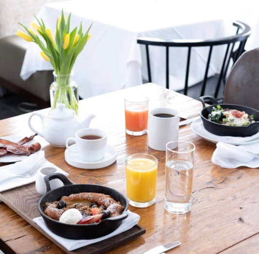 A wooden table set for breakfast with two small skillets of food, a teapot, cups of tea and coffee, orange juice, water, bacon, and a vase of yellow tulips—perfect for a bright and inviting Mother's Day Brunch in Stowe.