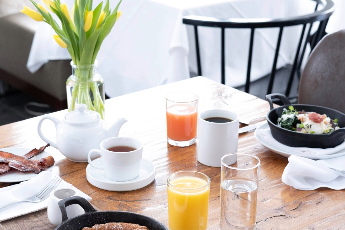 A breakfast table set with a teapot, cups of tea and coffee, glasses of orange and pink juice, bacon, oatmeal, pancakes, and a vase of yellow tulips—perfect for guests enjoying their morning as part of our vacation packages.