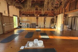 A serene yoga studio with wooden floors, sunlight streaming in, and yoga mats arranged in a circle. Crystal singing bowls sit on a rug beneath exposed wood beams draped with moss. Now featuring Reservation Enhancements for a seamless experience.