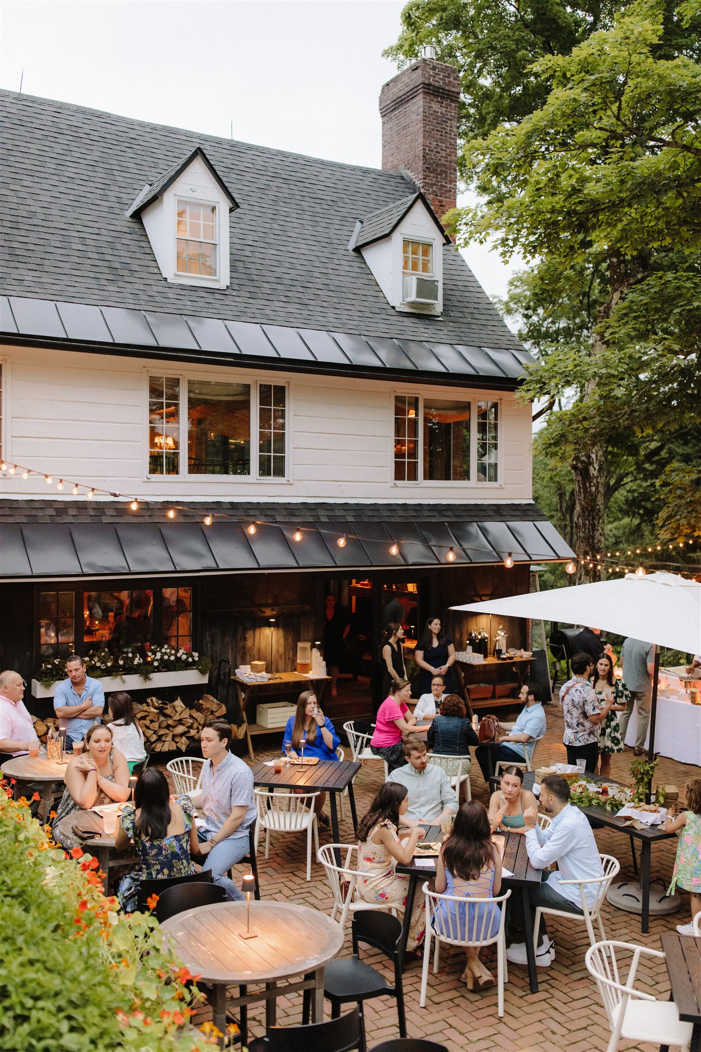 People dine outdoors at a restaurant patio under string lights, surrounded by greenery and a large house. The scene feels lively and social, with groups enjoying food and conversation.