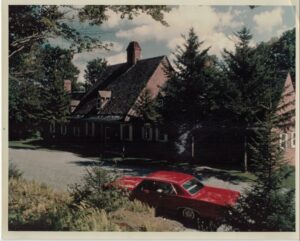 A red car is parked on a gravel driveway in front of a large brick house with a steep roof, surrounded by tall evergreen trees under a partly cloudy sky—the perfect setting for a relaxing Vermont vacation.