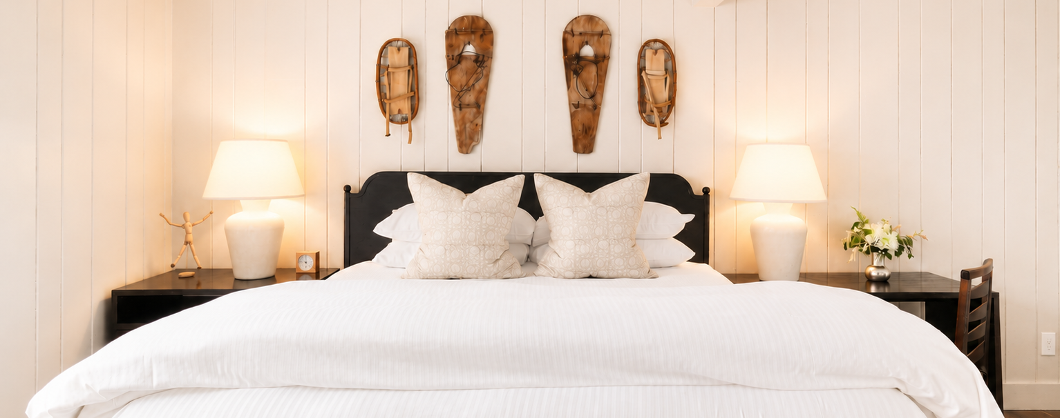 A cozy bedroom in one of the Most Romantic Hotels in Vermont, with a large bed covered in white linens, two bedside tables with lamps, and three rustic snowshoes mounted on the white paneled wall above the headboard. A cozy bedroom in one of the Most Romantic Hotels in Vermont, with a large bed covered in white linens, two bedside tables with lamps, and three rustic snowshoes mounted on the white paneled wall above the headboard.