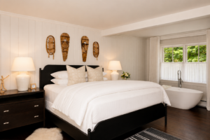 A cozy bedroom at one of the Most Romantic Hotels in Vermont, featuring a large bed with white linens, wooden masks above, and a freestanding bathtub by a bright window with flowing curtains.