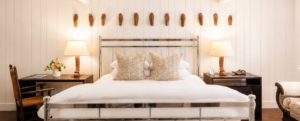 Bright bedroom with a metal-frame bed, patterned pillows, matching dark nightstands and lamps, a desk with a chair, and wall-mounted masks above the bed. White paneled walls and airy decor create the cozy, inviting feel reminiscent of a Vermont luxury hotel.
