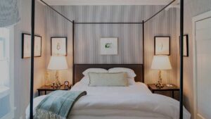 A neatly made bed with white linens and a green throw blanket sits between two lamps on nightstands, creating the inviting atmosphere characteristic of Edson Hill Guest Rooms, with framed art on the striped gray walls and a window to the left.