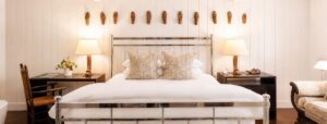 A stylish bedroom reminiscent of a Vermont luxury hotel, featuring a metal-frame bed, two patterned pillows, lamps on dark nightstands, a wooden chair, and decorative items on the wall above the bed. The room showcases white paneled walls and warm lighting for an inviting ambiance.