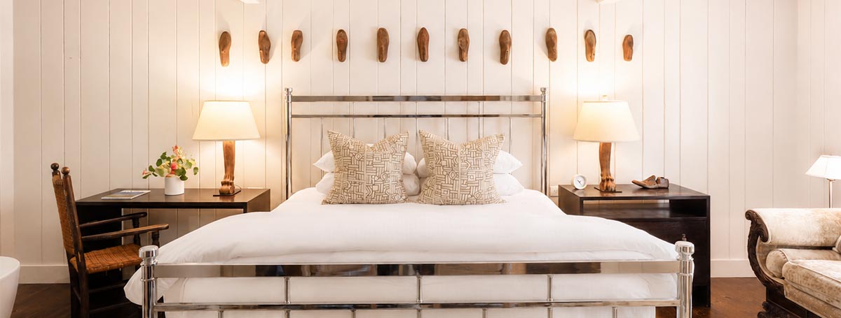 A stylish bedroom reminiscent of a Vermont luxury hotel, featuring a metal-frame bed, two patterned pillows, lamps on dark nightstands, a wooden chair, and decorative items on the wall above the bed. The room showcases white paneled walls and warm lighting for an inviting ambiance. A stylish bedroom reminiscent of a Vermont luxury hotel, featuring a metal-frame bed, two patterned pillows, lamps on dark nightstands, a wooden chair, and decorative items on the wall above the bed. The room showcases white paneled walls and warm lighting for an inviting ambiance.