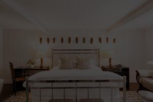 Bright bedroom at Edson Hill with a metal-framed bed, patterned pillows, two bedside tables with lamps, wooden chair, and several masks or decor pieces mounted on the white-paneled wall above the bed.