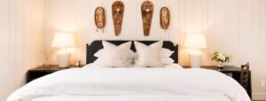 A neatly made bed with white linens, two large pillows, and bedside tables with lamps sets a cozy scene reminiscent of the Most Romantic Hotels in Vermont. Three wooden wall decorations hang above the bed, and a chair and small vase of flowers are placed on the right side, adding to the inviting ambiance.