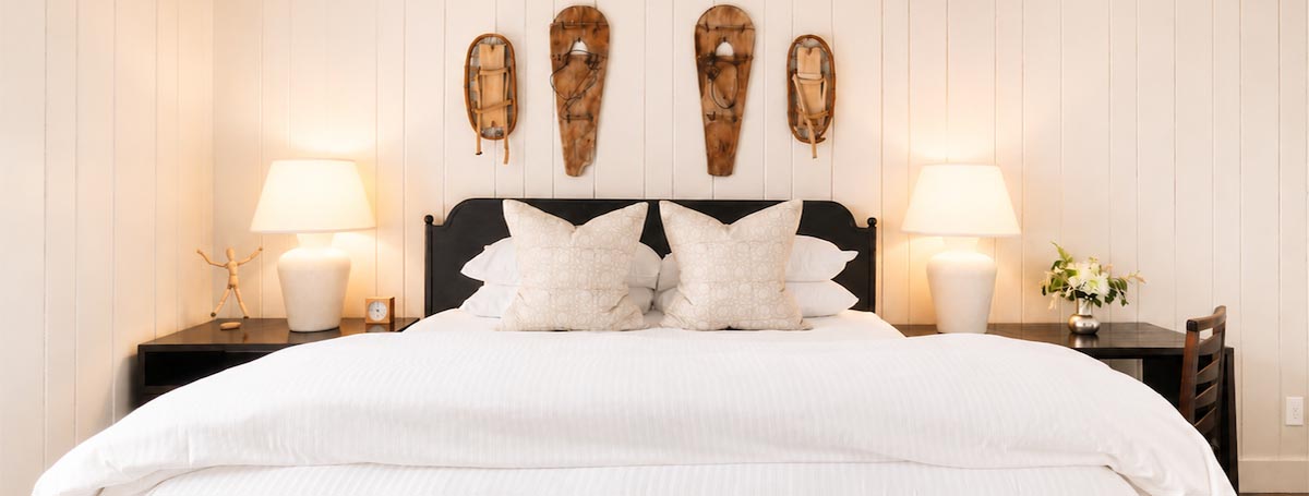 A neatly made bed with white linens, two large pillows, and bedside tables with lamps sets a cozy scene reminiscent of the Most Romantic Hotels in Vermont. Three wooden wall decorations hang above the bed, and a chair and small vase of flowers are placed on the right side, adding to the inviting ambiance. A neatly made bed with white linens, two large pillows, and bedside tables with lamps sets a cozy scene reminiscent of the Most Romantic Hotels in Vermont. Three wooden wall decorations hang above the bed, and a chair and small vase of flowers are placed on the right side, adding to the inviting ambiance.