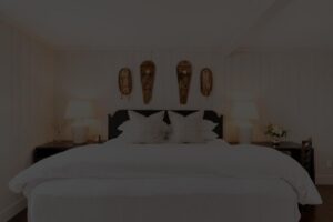 A neatly made bed with white bedding, two large pillows, lamps on bedside tables, and three decorative masks hanging on the wall above the headboard creates an inviting atmosphere reminiscent of Edson Hill’s cozy elegance.