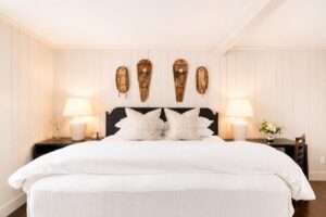 A cozy bedroom inspired by Edson Hill features a large white bed with decorative pillows, two bedside tables with lamps, wall-mounted snowshoes above the headboard, and a small vase of flowers on the right table.