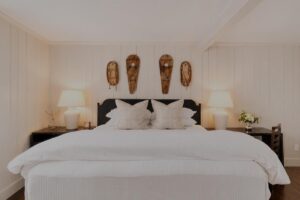 A neatly made bed with white linens, two large pillows, and two lamps on dark bedside tables evokes the tranquil charm of Edson Hill. Three rustic wooden decorations hang on the white paneled wall above the headboard, adding a touch of warmth to this inviting setting.