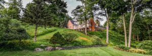 A large brick house sits atop a grassy hill surrounded by tall trees, manicured shrubs, rocks, and green gardens on a sunny day—offering the timeless charm reminiscent of Edson Hill - Stowe Vermont Inn.