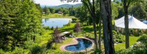 A heart-shaped pool, lounge chairs, and umbrellas sit amid lush greenery near a small lake at Edson Hill - Stowe Vermont Inn, with a white tent to the right and rolling hills in the background under a clear sky.