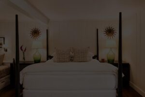 A neatly made bed with white linens and multiple pillows, flanked by green lamps on dark nightstands, evokes the cozy elegance of Edson Hill. Two sunburst wall decorations hang above the bed, and a window seat is visible on the left side.