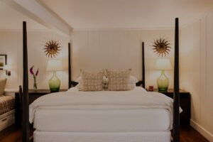 A neatly made bed with white bedding and patterned pillows is centered between two nightstands, each with a green lamp and sunburst wall decor above them, creating an inviting ambiance reminiscent of Edson Hill in this cozy, well-lit bedroom.