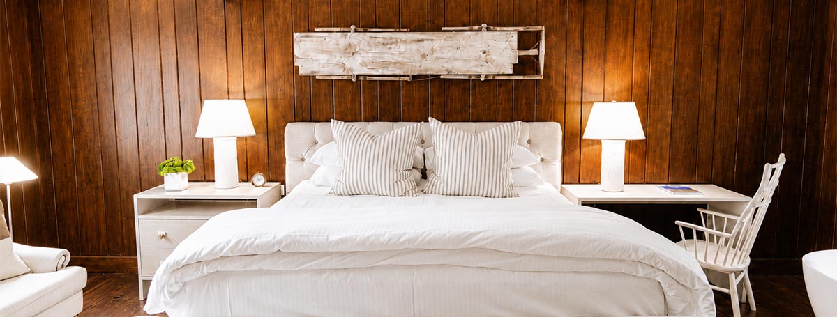 A cozy bedroom at Edson Hill features a white bed, striped pillows, two bedside tables with lamps, a white chair, and wooden paneled walls. A decorative wooden piece hangs above the bed. A cozy bedroom at Edson Hill features a white bed, striped pillows, two bedside tables with lamps, a white chair, and wooden paneled walls. A decorative wooden piece hangs above the bed.