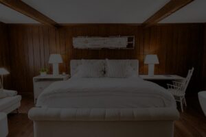 A cozy bedroom at Edson Hill with wood-paneled walls, a large bed with white bedding, two bedside tables with lamps, a white armchair, and a white chair at a desk.
