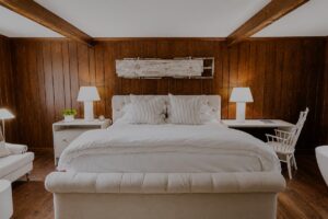 A cozy bedroom at Edson Hill features wood-paneled walls, a white upholstered bed with striped pillows, two white nightstands with lamps, and a white chair at a desk. Exposed ceiling beams add rustic charm.