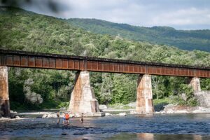 A rusty railroad bridge spans over a shallow river with four people standing on rocks below, enjoying a watersports experience surrounded by dense green forest and hills in the background.